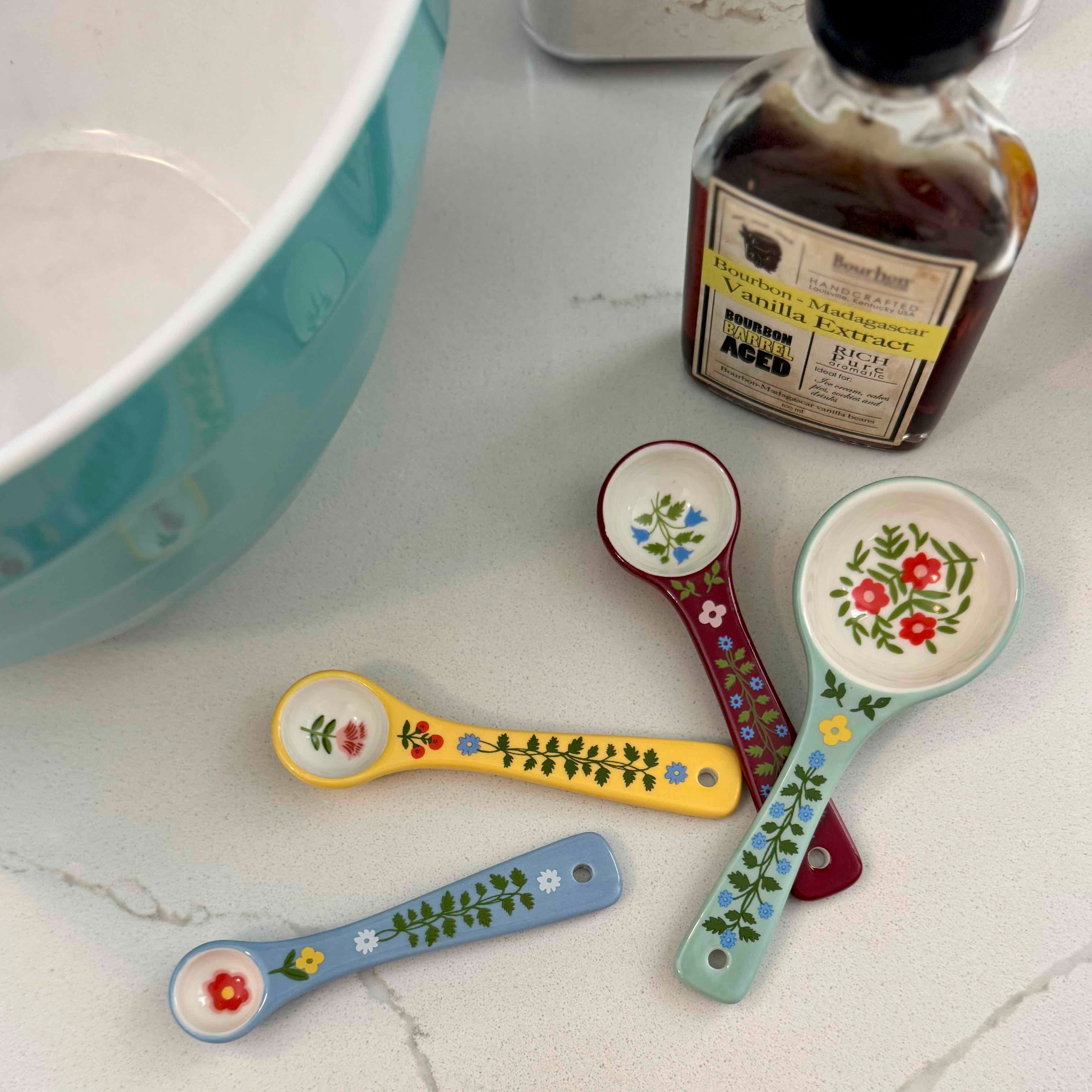 Colorful ceramic measuring spoons with floral designs on a marble surface next to a bottle of vanilla extract.