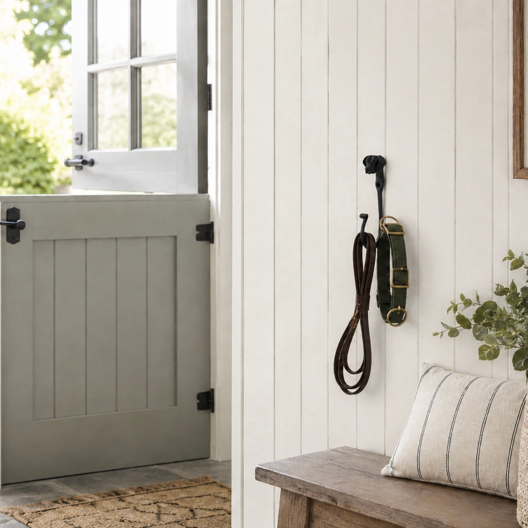 A rustic interior scene with a white shiplap wall holding black wall hooks, one with a coiled brown strap and one with a green and gold strap. A gray barn-style door is open on the left, and a wooden bench with a striped pillow sits on the right.