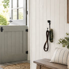 A rustic interior scene with a white shiplap wall holding black wall hooks, one with a coiled brown strap and one with a green and gold strap. A gray barn-style door is open on the left, and a wooden bench with a striped pillow sits on the right.