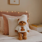 Teddy bear in a white outfit with a teddy bear design, sitting on a bed with pillows and fairy lights in the background.