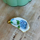 A small teardrop-shaped white dish with a gold-trimmed edge, decorated with blue flowers and green leaves, resting on a light wood surface.