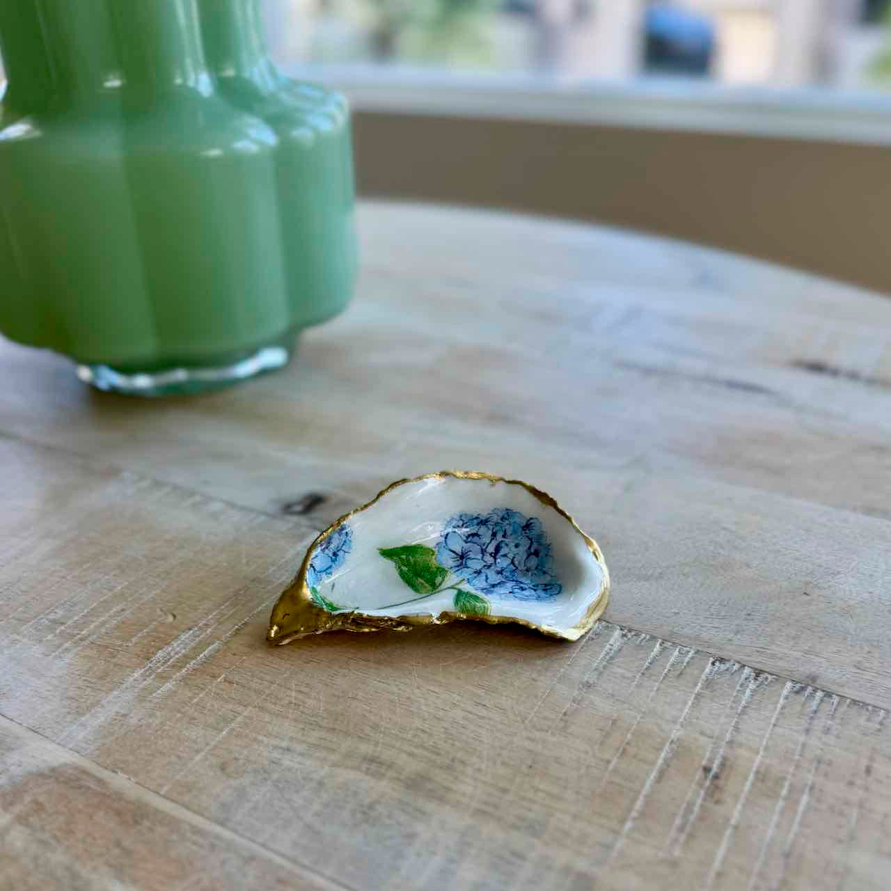 A small decorative oyster shell dish with hand-painted blue flowers, green leaves, and a gold rim, resting on a light wood tabletop next to a blurred mint green vase.