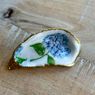 A hand-painted resin or glass dish shaped like an oyster shell, featuring blue hydrangea flowers, green leaves, and a gold-rimmed edge, resting on a light wood surface.