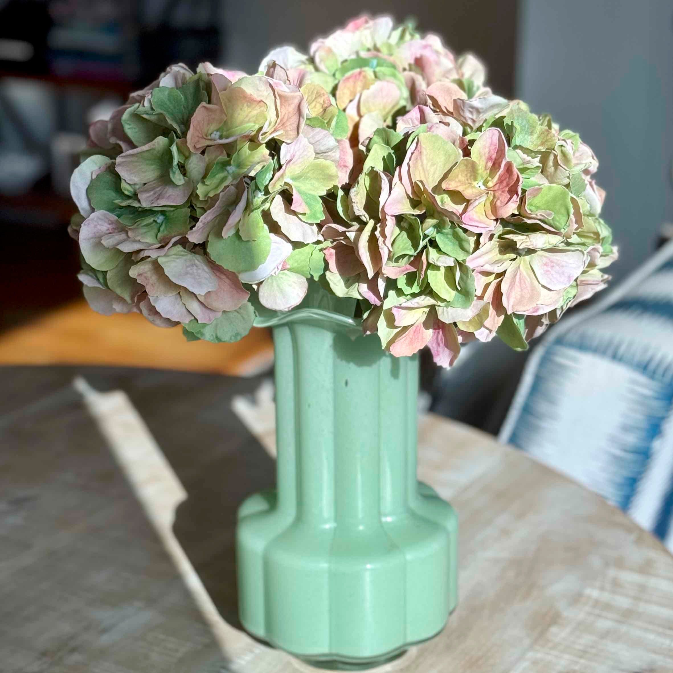 A pale mint green ceramic vase holding a large bouquet of faded pink and green hydrangea flowers, placed on a wooden table in a softly lit indoor setting.
