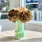 A pale mint green ribbed glass vase holding a bouquet of dried pink and olive hydrangea flowers, set on a light wood table in front of a softly blurred window.