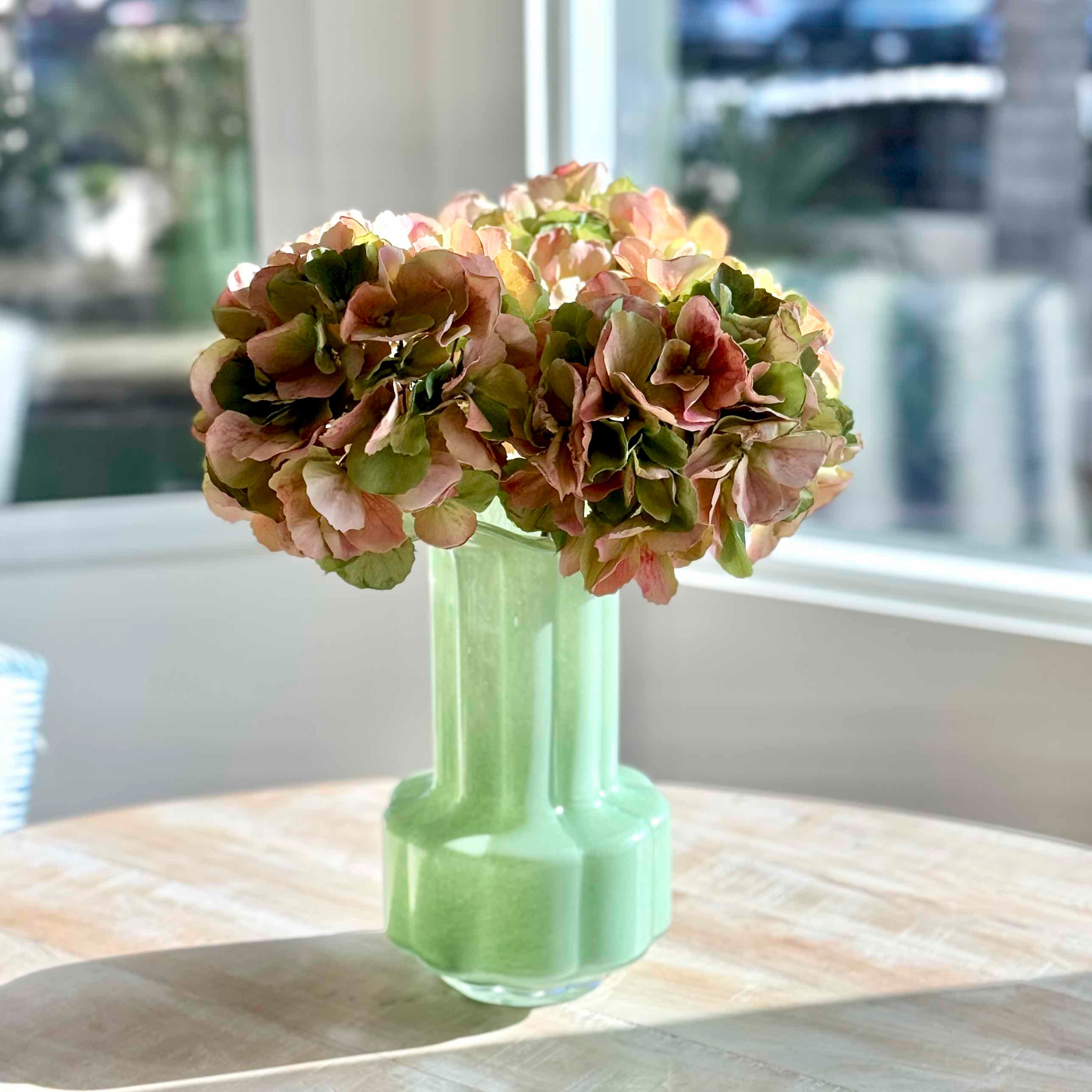 A pale mint green ribbed glass vase holding a bouquet of dried pink and olive hydrangea flowers, set on a light wood table in front of a softly blurred window.