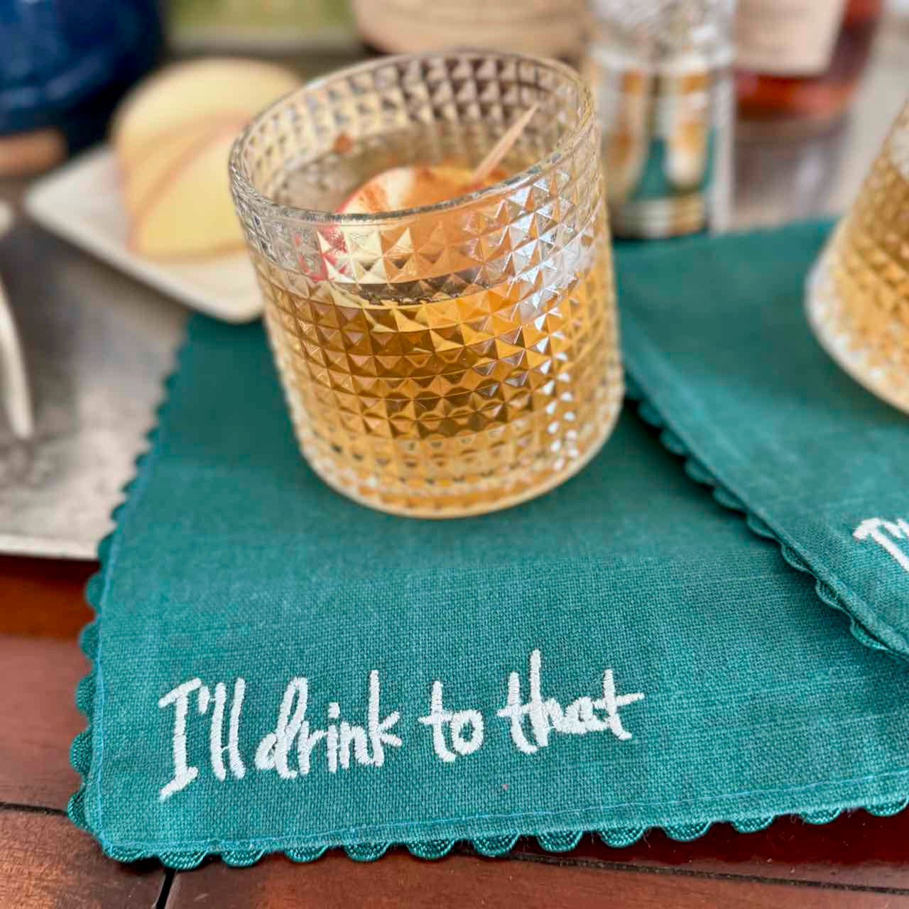 Teal napkin with 'I'll drink to that' text on a wooden table with a glass of whiskey.