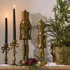 Two gold-toned nutcracker figurines displayed on a tabletop beside black taper candles and a green evergreen plant, with a warm indoor background.