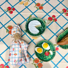 A styled flat-lay of a matching tablecloth featuring a blue grid pattern and red cherries, accompanied by coordinating ceramic dishes, a patterned napkin, and bread on a table setting.