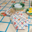 A folded cloth napkin with a cherry print on a cream background, tied with a clear plastic fastener and placed on a patterned tablecloth. In the background, there are small dishes with food items.