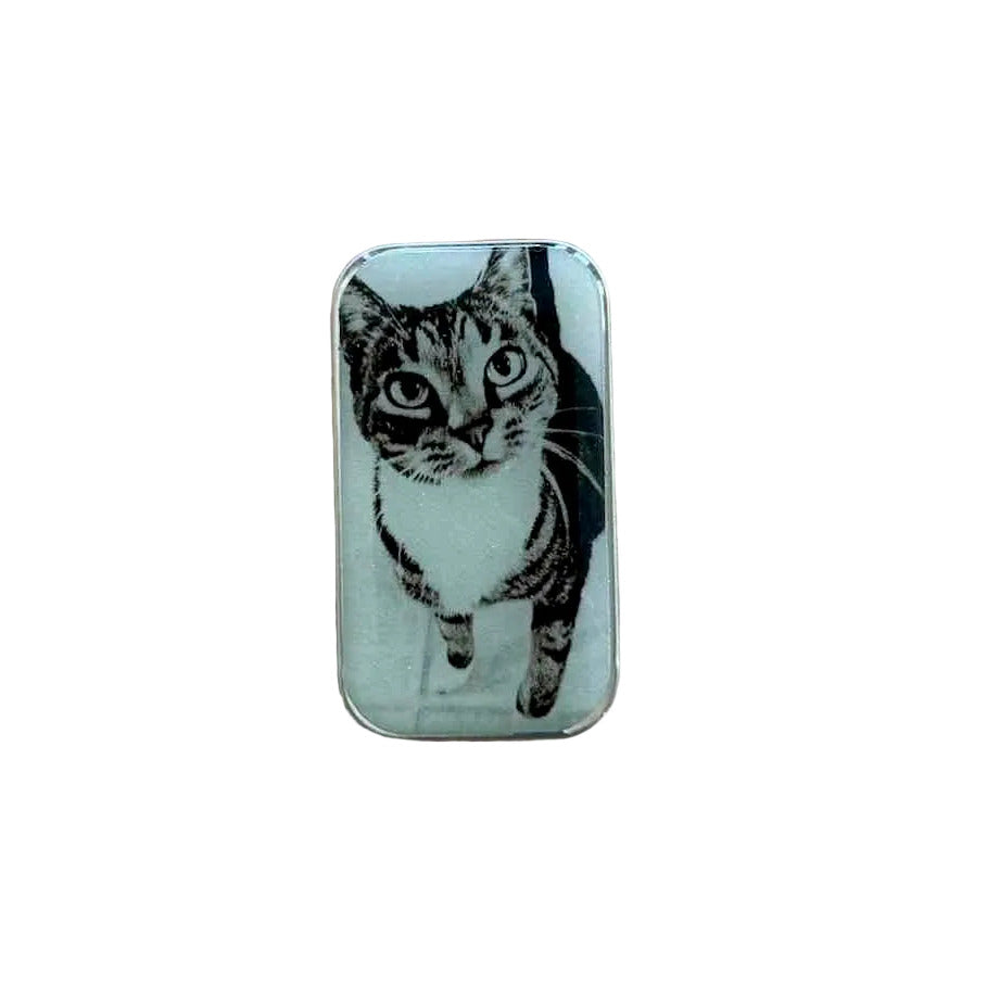 A rectangular metal pendant with rounded corners featuring a black and white illustration of a cat, resting on a light-colored wooden surface.