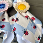 A close-up of a light blue and white checkered napkin with a repeating pattern of red and black ladybugs, decorated with two flower-shaped napkin rings, one yellow and one pink.