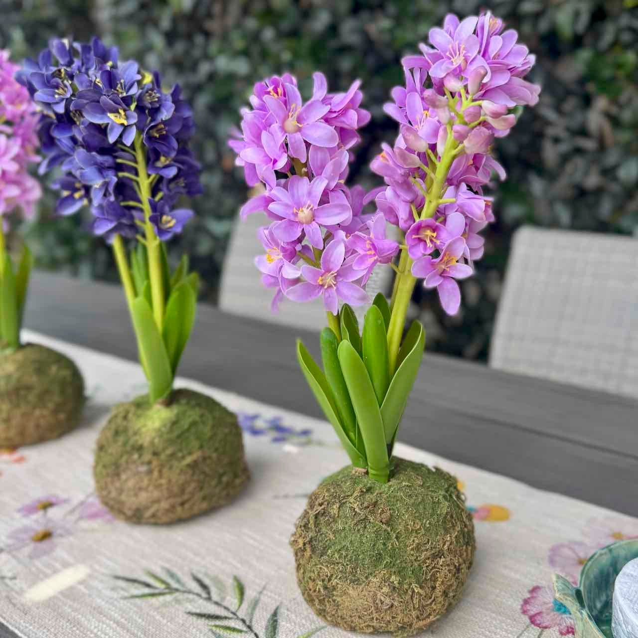 Three artificial hyacinth table centerpieces with purple and blue flowers in moss balls, arranged on a floral table runner.