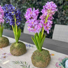 Three artificial hyacinth table centerpieces with purple and blue flowers in moss balls, arranged on a floral table runner.