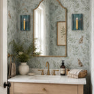 A styled bathroom vanity featuring a large arched mirror with a gold frame, set against a light blue-green botanical wallpaper with rabbit motifs. The vanity has a white marble sink top, brass fixtures, and is decorated with a ceramic vase of greenery, a folded white towel, and a wicker basket.