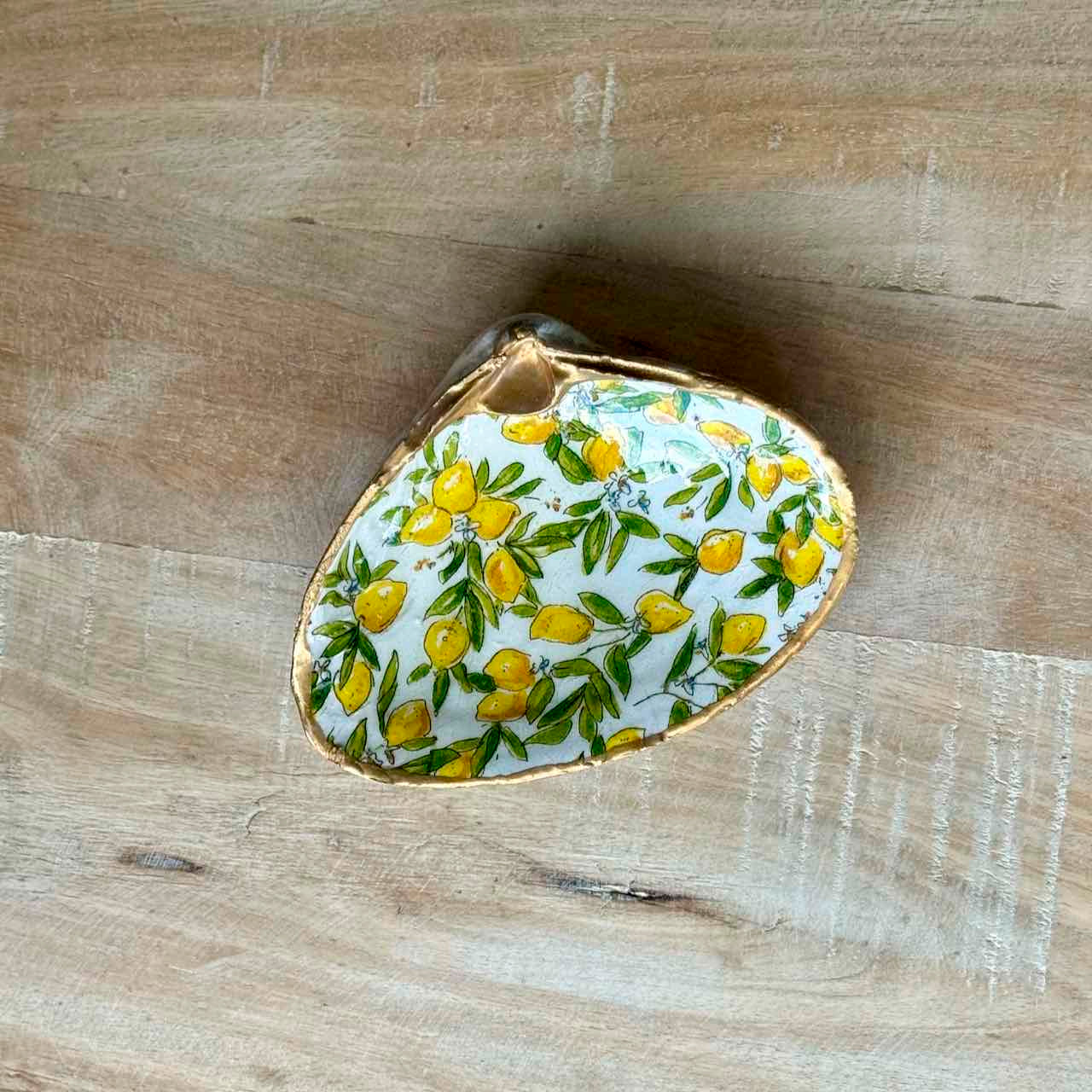 A small decorative shell-shaped dish with a white background, hand-painted yellow lemons, green leaves, and a gold rim, placed on a light wood surface.