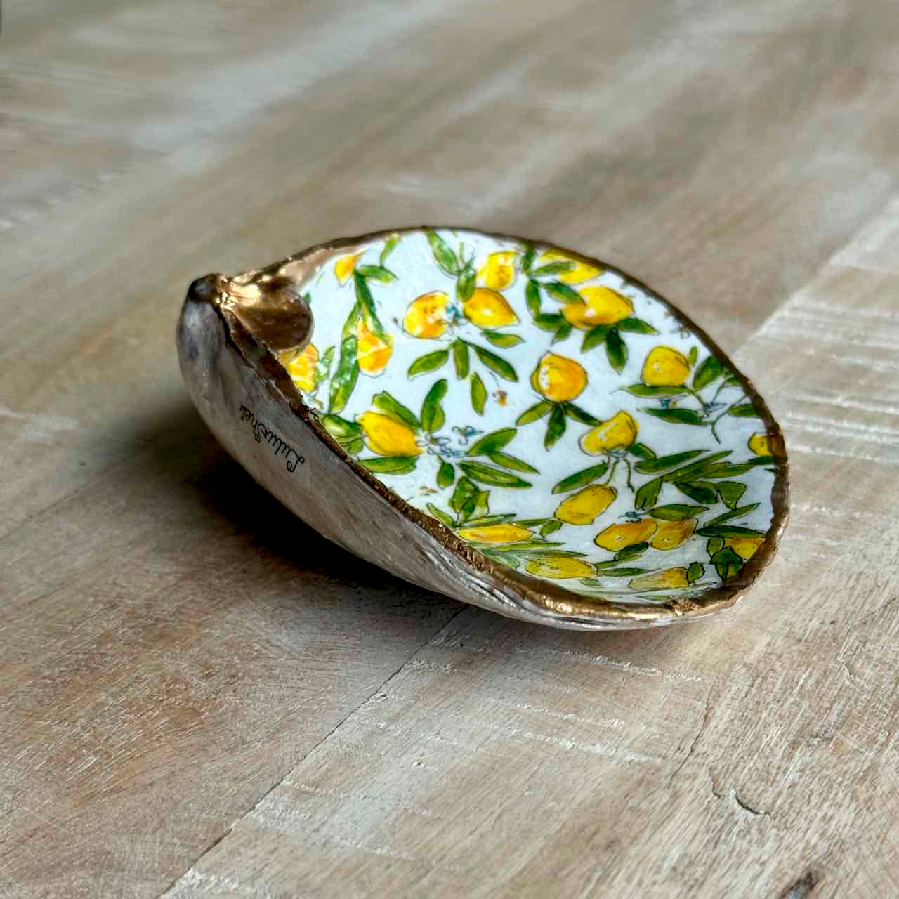 A small decorative dish shaped like a half shell, featuring a white interior printed with yellow lemons and green leaves, with a metallic gold-toned rim, placed on a light wood surface.