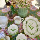 A collection of vintage ceramic tableware, including plates, a creamer, and a duck-shaped pitcher, arranged on a colorful floral tablecloth alongside a vase of pink hydrangeas.