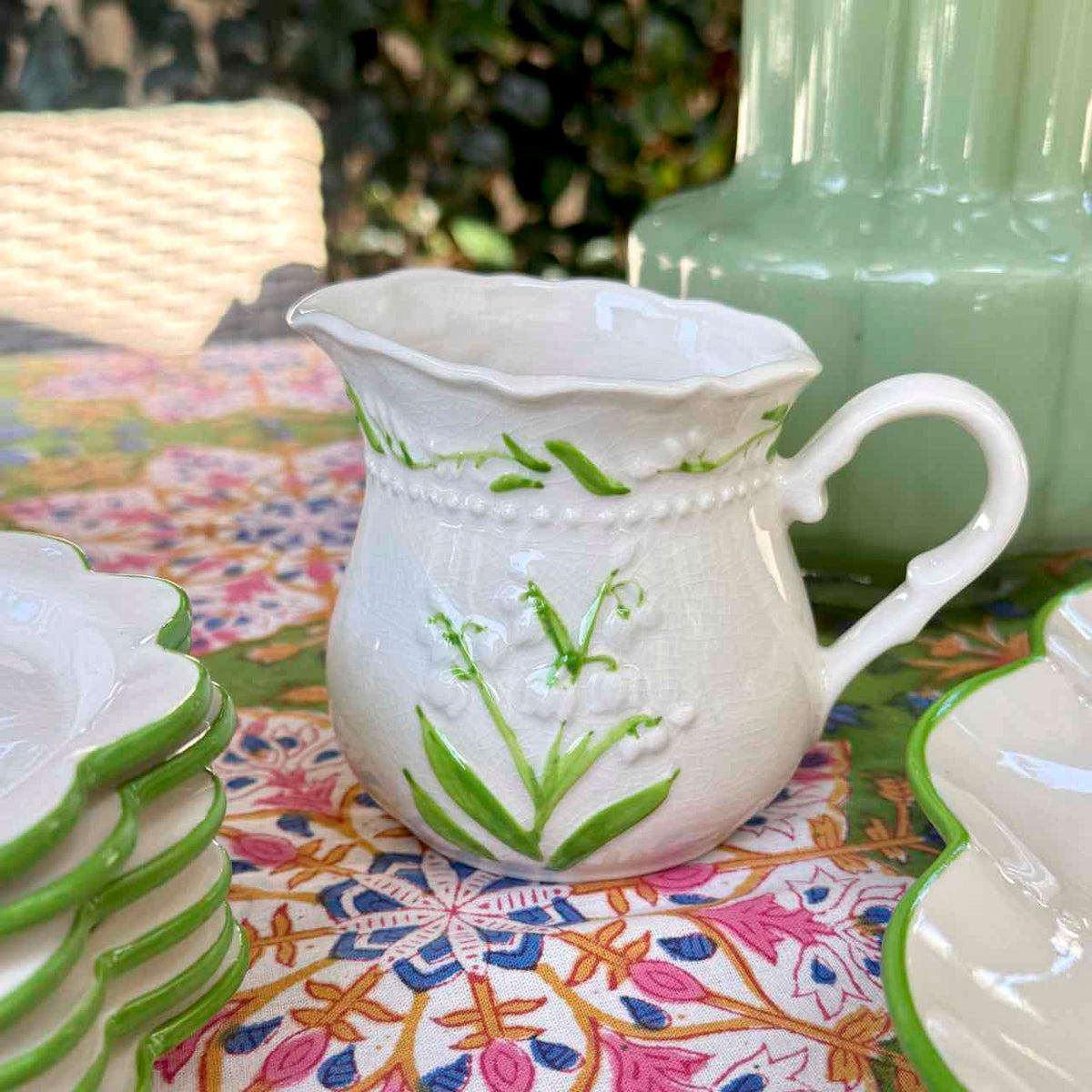 A white ceramic creamer pitcher decorated with green lily of the valley flowers and leaves, set on a colorful patterned tablecloth outdoors.