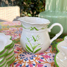 A white ceramic creamer pitcher decorated with green lily of the valley flowers and leaves, set on a colorful patterned tablecloth outdoors.