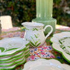 A white ceramic pitcher and matching plates with green leaf and floral patterns, arranged on a colorful floral tablecloth with a green glass vase in the background.