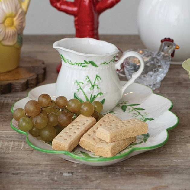 A white ceramic serving pitcher with green floral vine patterns sits on a matching scalloped plate with green edging, styled with green grapes and rectangular biscuits on a rustic wooden table. A small clear glass chicken figurine and other decorative objects are blurred in the background.