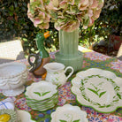 A styled collection of ceramic tableware displayed outdoors on a floral tablecloth, featuring a green duck-shaped pitcher, a ribbed light green vase with pink hydrangea flowers, a white embossed bowl, a cream pitcher with a green handle, and multiple white plates and bowls decorated with green floral and lily-of-the-valley motifs.