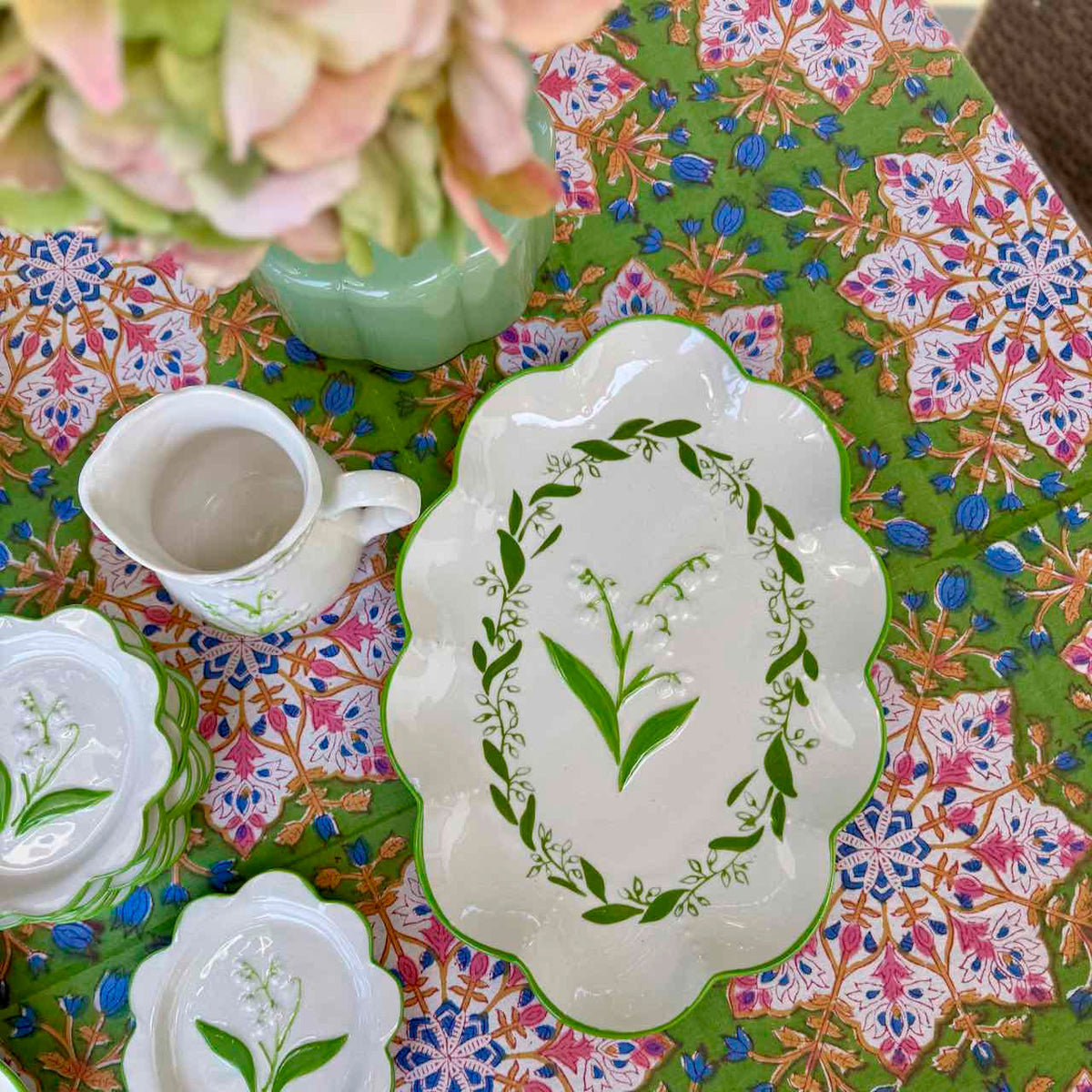 A white ceramic floral serving platter with scalloped edges and a green lily of the valley leaf design, styled on a colorful patterned tablecloth with matching dishes and a vase of flowers.