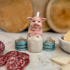 Ceramic pig candle with small barrels on a marble surface with cured meats and bread in the background.