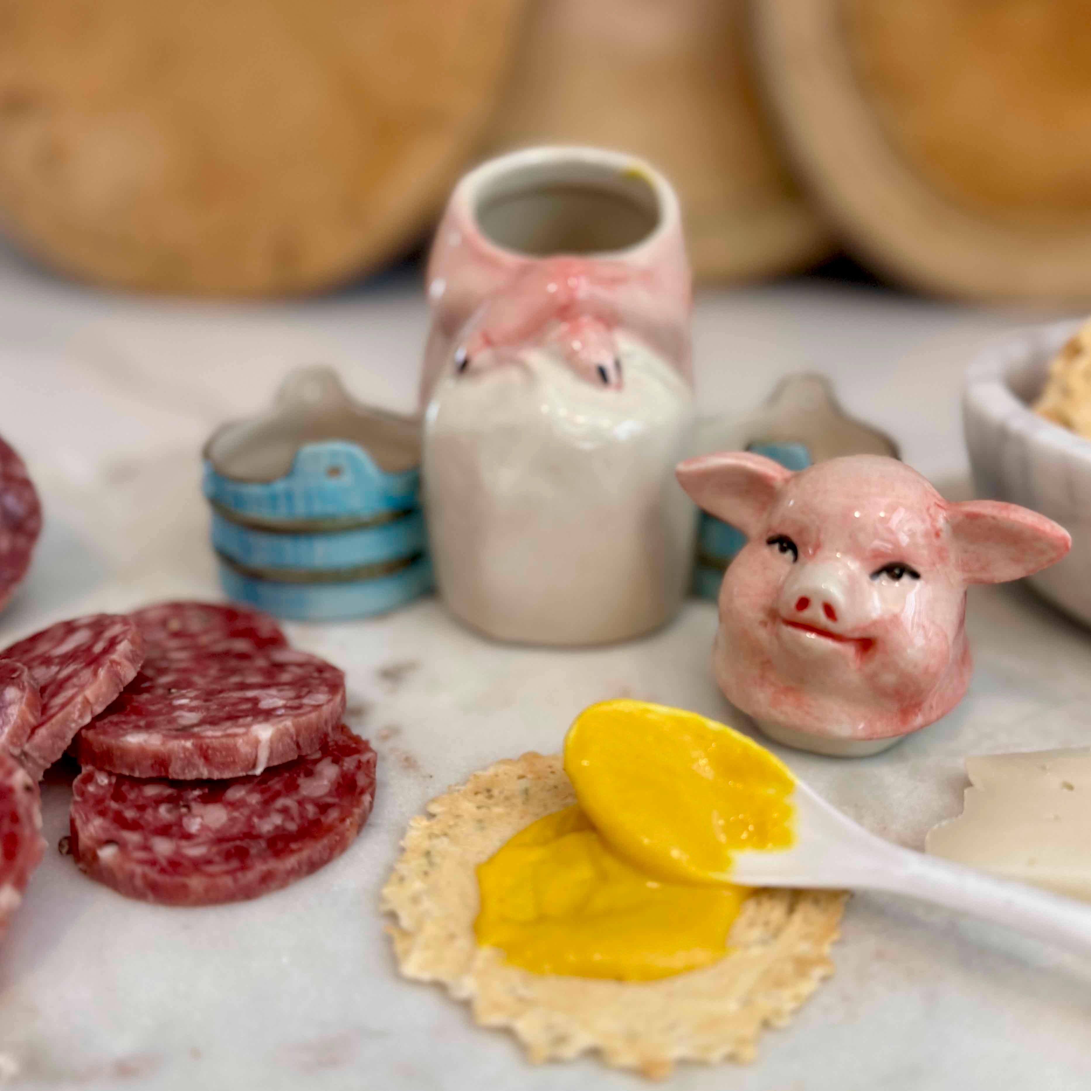 Ceramic pig head with mustard on a cracker, surrounded by salami and a jar of mustard.