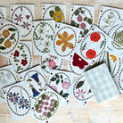 A spread of floral playing cards arranged on a light wood surface, with one card showing a white-and-gray plaid back. The cards feature pressed flower illustrations in many colors and suit symbols in the corners.