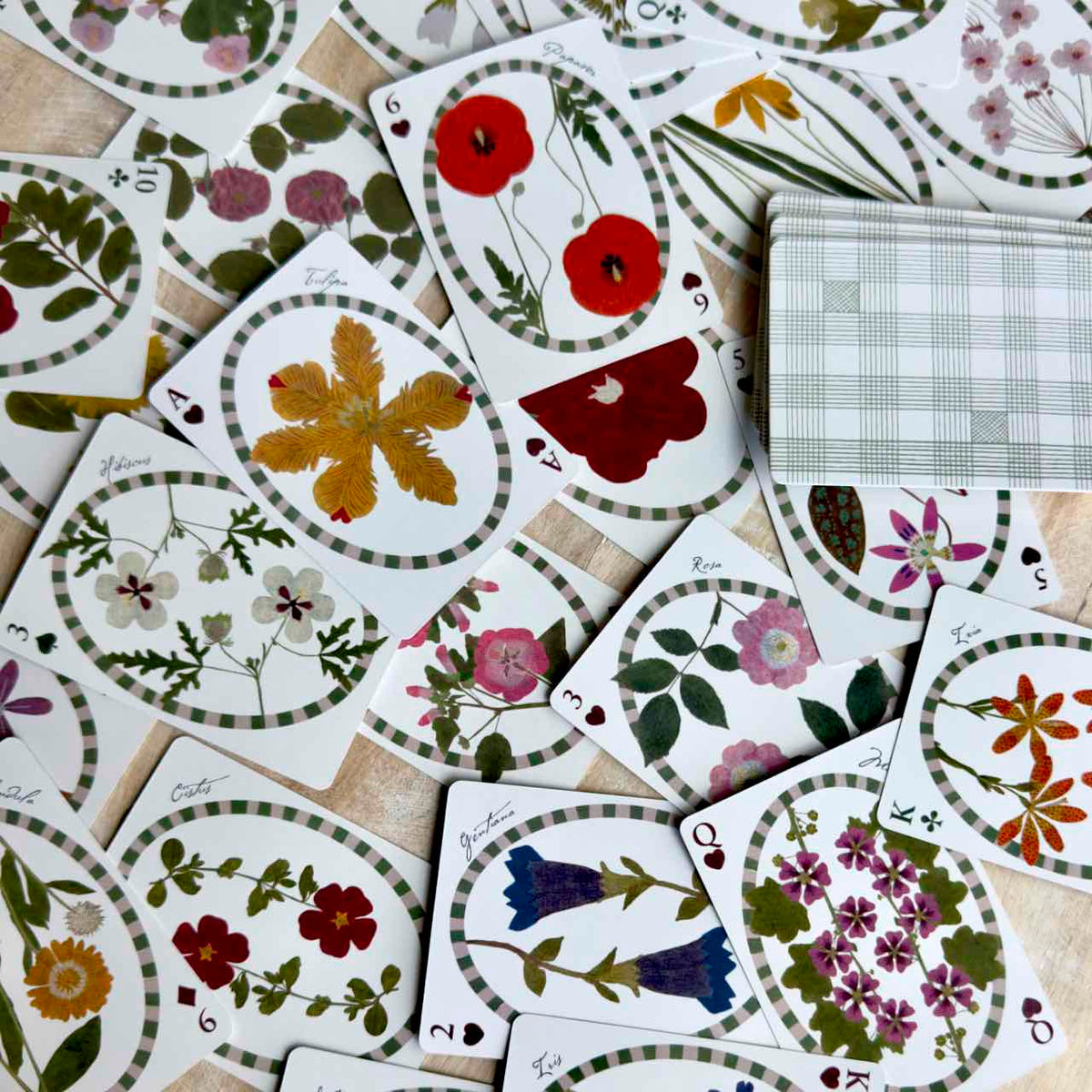 A spread of decorative playing cards featuring pressed floral illustrations, with a stack of cards showing a green-and-white plaid pattern on a light wood surface.