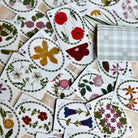 A spread of decorative playing cards featuring pressed floral illustrations, with a stack of cards showing a green-and-white plaid pattern on a light wood surface.
