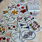 A spread of illustrated playing cards on a light wood surface, with a small stack of cards showing a white-and-green plaid back. The cards feature oval floral and botanical designs in many colors including red, pink, blue, yellow, green, and white.
