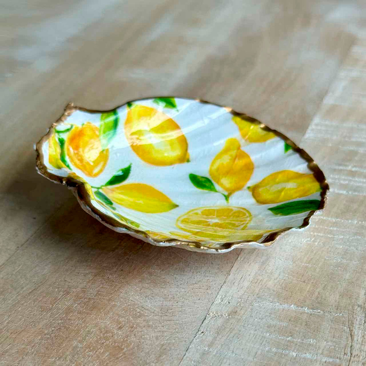 A small decorative dish shaped like a clamshell, featuring a hand-painted lemon and leaf pattern on a white background with a gold-toned rim, set on a light wood surface.