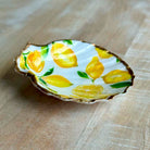 A small decorative dish shaped like a clamshell, featuring a hand-painted lemon and leaf pattern on a white background with a gold-toned rim, set on a light wood surface.