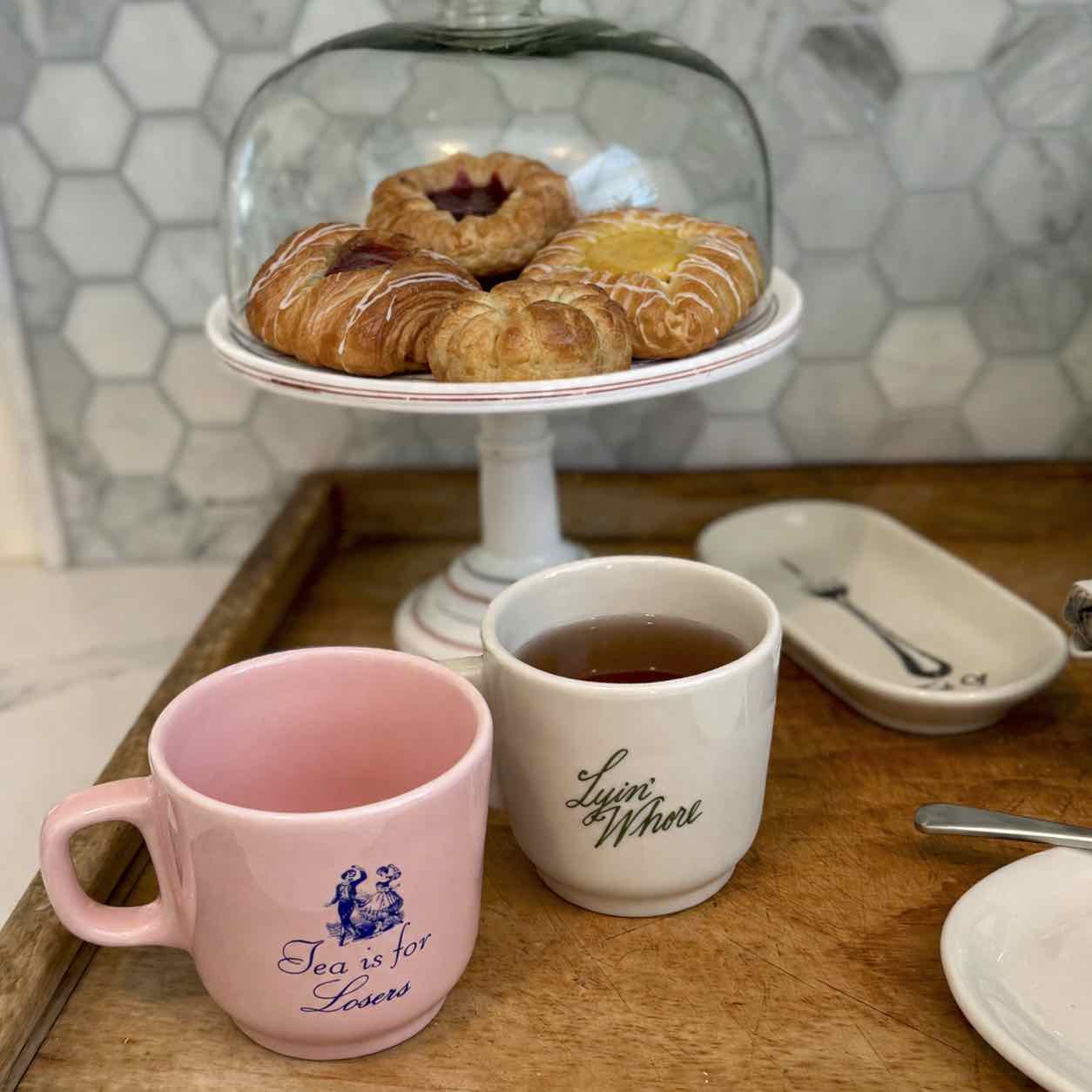 Two ceramic mugs on a wooden tray in a kitchen setting, with a pink mug reading 'She is for Lovers' and a white mug reading 'Love is Whate'. A glass-domed pastry stand with pastries sits behind them, and a tiled wall is visible in the background.