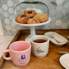 Two ceramic mugs on a wooden tray in a kitchen setting, with a pink mug reading 'She is for Lovers' and a white mug reading 'Love is Whate'. A glass-domed pastry stand with pastries sits behind them, and a tiled wall is visible in the background.