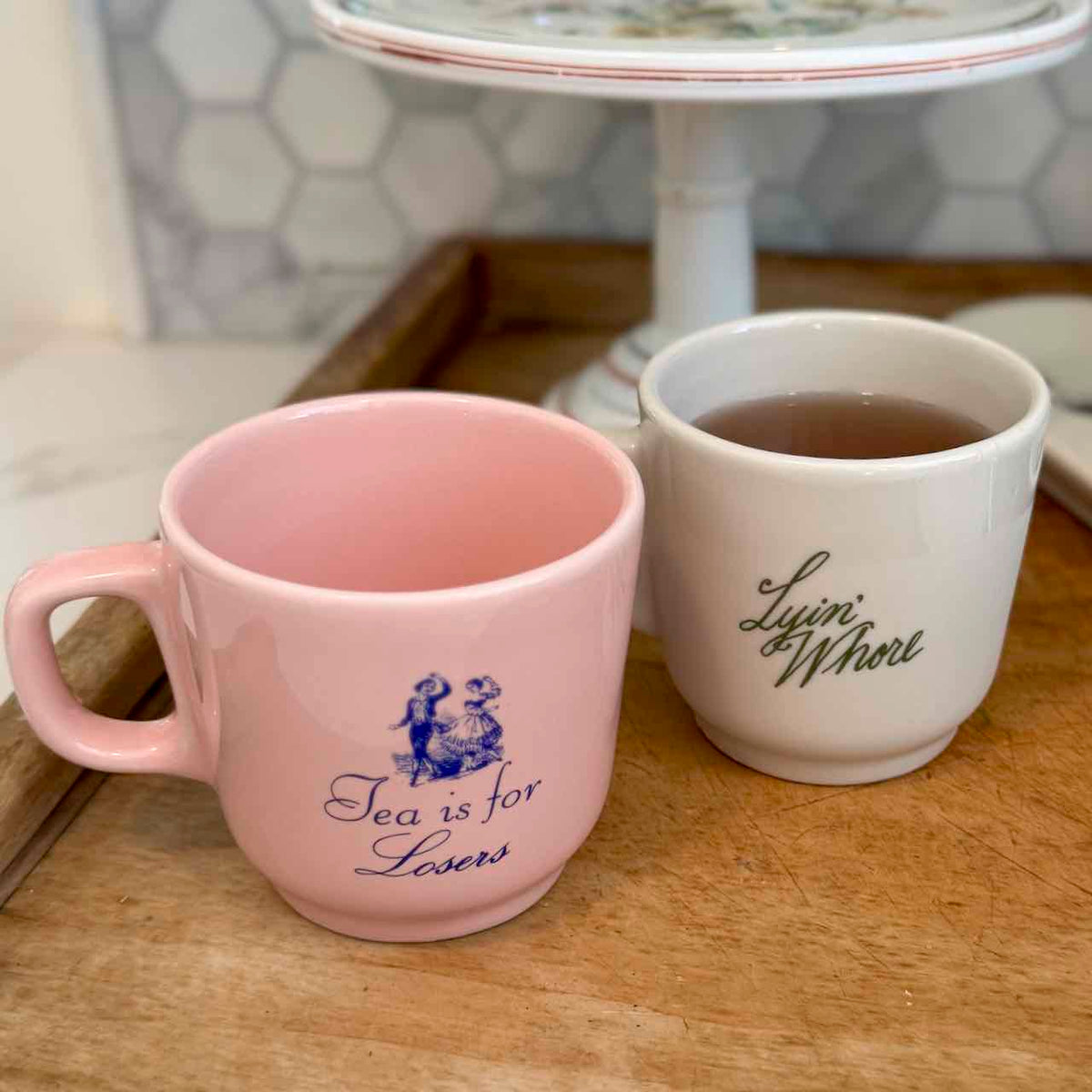Two ceramic tea mugs on a wooden table, one light pink with blue text reading 'Tea is for losers' and one white with green script reading 'Lynni's Whirl'.