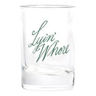 A clear glass rocks tumbler with the phrase 'Lyin' Whore' printed in dark green cursive script on the front, set against a plain white background.