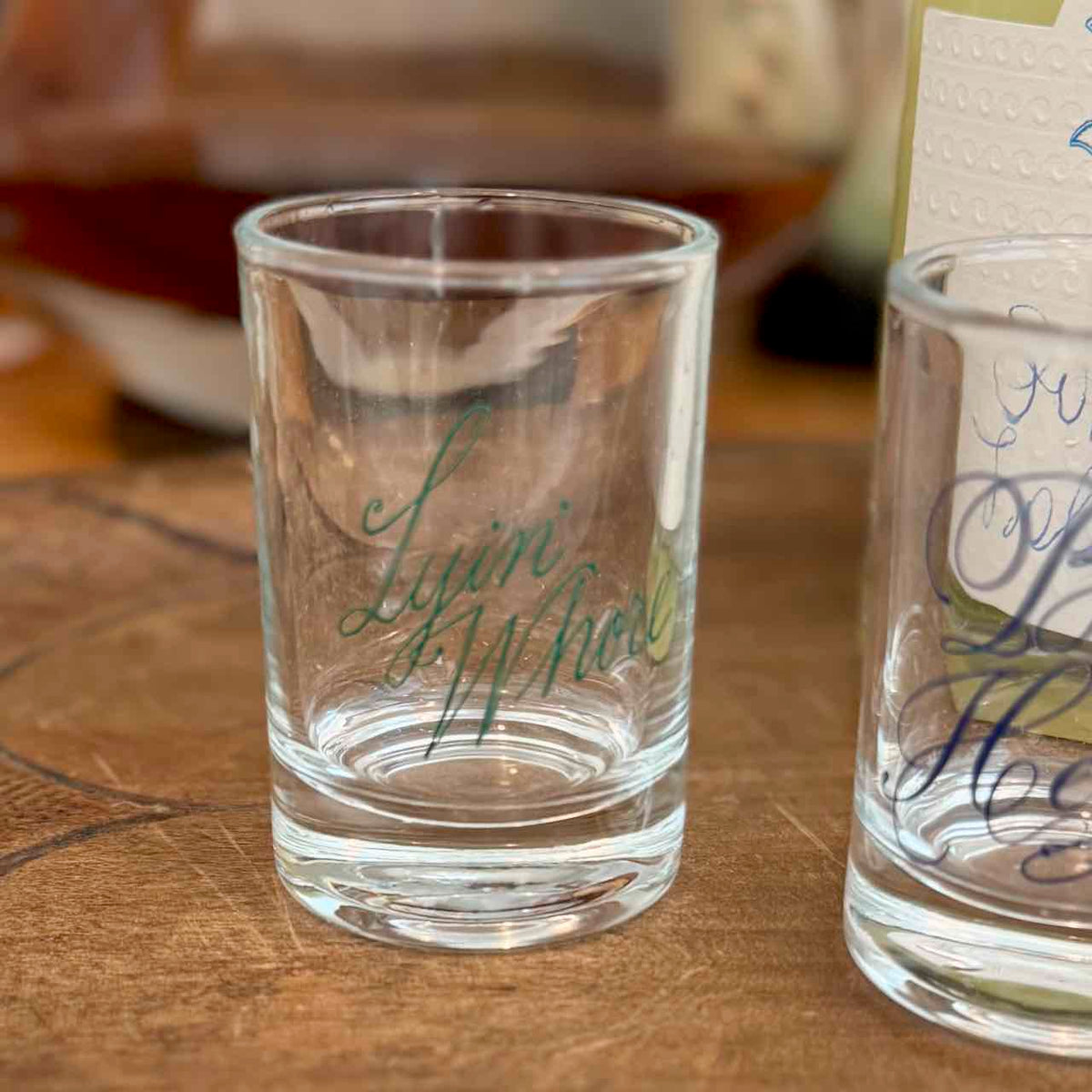 A clear glass shot glass with the name 'Lyli Whate' written in green cursive script, sitting on a wooden tabletop. Another partially visible shot glass with blue script is on the right, and a blurred indoor background is behind.