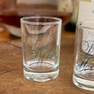A clear glass shot glass with the name 'Lyli Whate' written in green cursive script, sitting on a wooden tabletop. Another partially visible shot glass with blue script is on the right, and a blurred indoor background is behind.