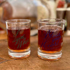 Two clear shot glasses filled with amber liquid sit on a wooden table. The left glass has green cursive text reading 'Lyin' Whoo', and the right glass has blue cursive text reading 'Bless your Heart'.