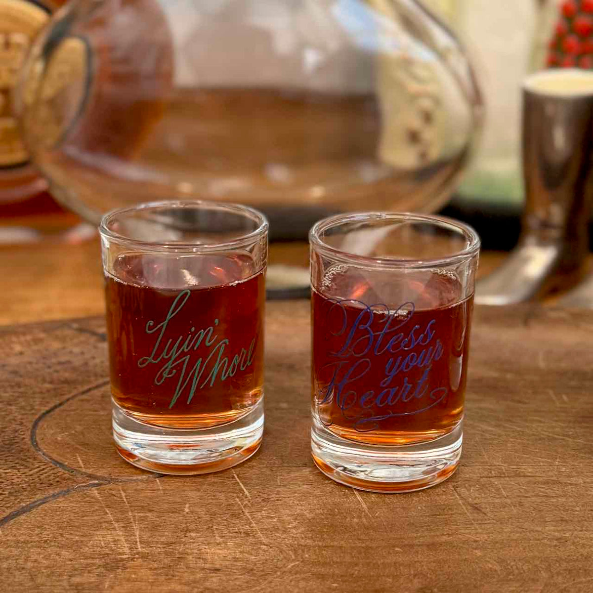 Two clear glass shot glasses filled with amber liquid sit on a wooden surface. The left glass is etched with the words 'Lyini Mowri' in green, and the right glass is etched with 'Bless your Heart' in blue.