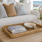 A rectangular woven rattan serving tray with handles sits on a light wood table in a bright living room. The tray holds a stack of books, a white bowl of decorative stones, and a tassel, with a white sofa and pillows behind it and a window view of the ocean.