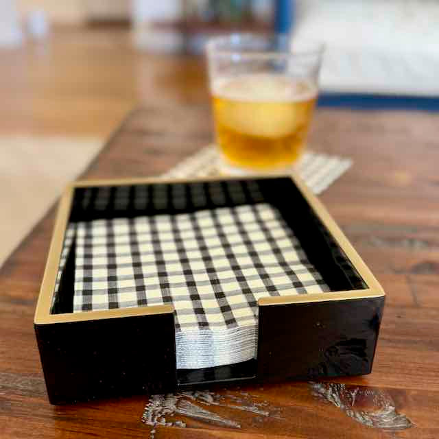 Black and gold square tray with checkered napkins on a wooden table with a glass of amber liquid in the background.