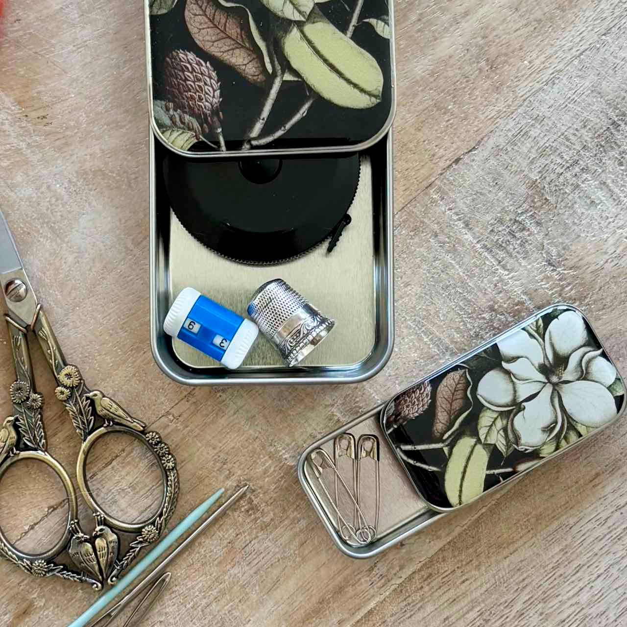 A set of decorative metal sewing and storage cases with a black floral and botanical print, shown on a light wood tabletop alongside vintage-style scissors, tweezers, and thimbles.