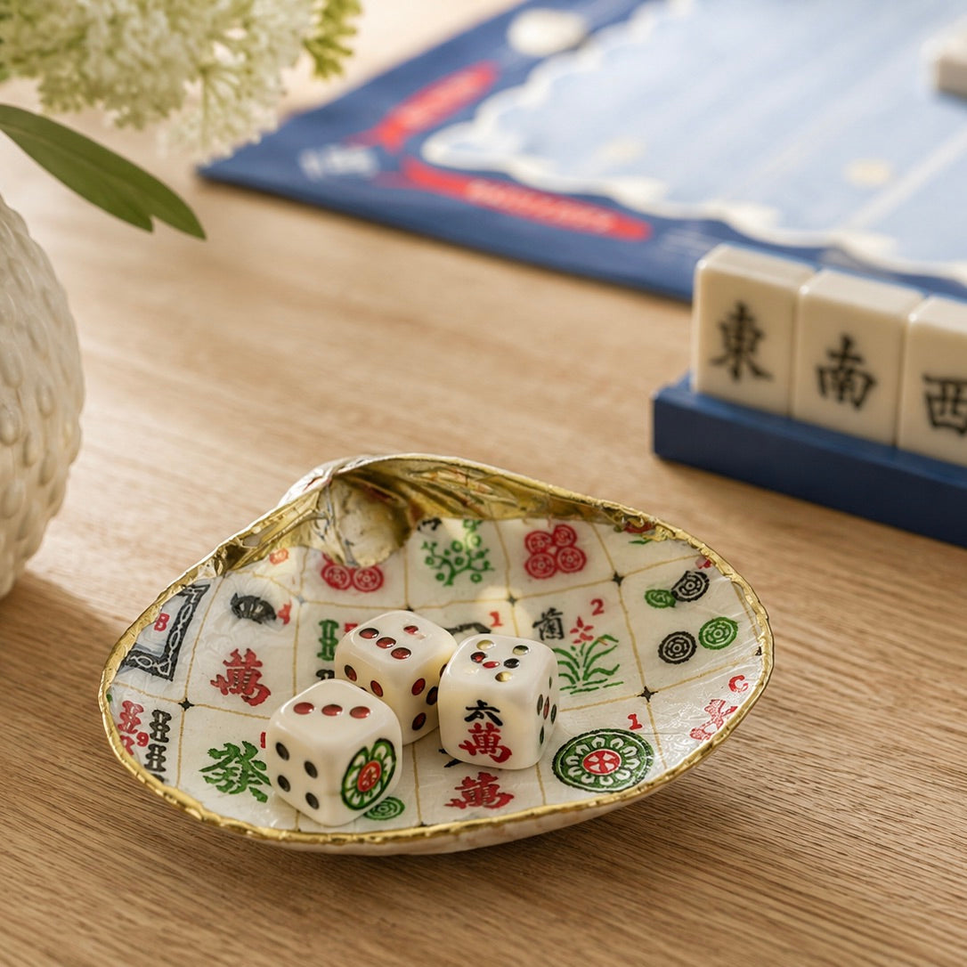 A decorative ceramic dish shaped like a shell, featuring a white and gold Mahjong tile pattern, holding three white dice with black markings on a wooden tabletop.