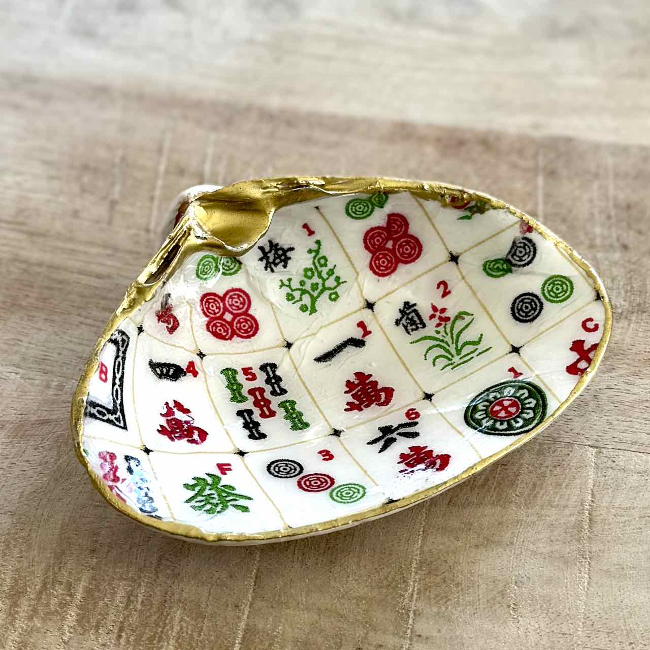 A decorative shell-shaped dish featuring a printed Mahjong tile pattern with gold trim, photographed on a light wood surface.