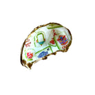 A handmade ring shaped like a broken shell, featuring a white enamel face decorated with colorful Chinese-style tiles, characters, and motifs, and finished with a gold-tone rim, resting on a light wood surface.
